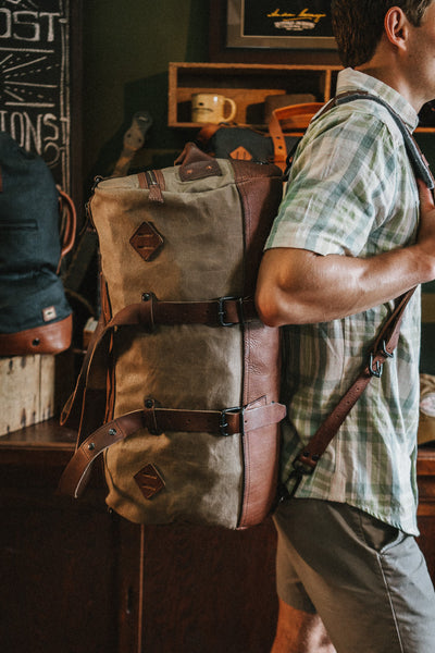 Brown leather straps of a backpack, secured with metal hardware, worn by someone in a plaid shirt.